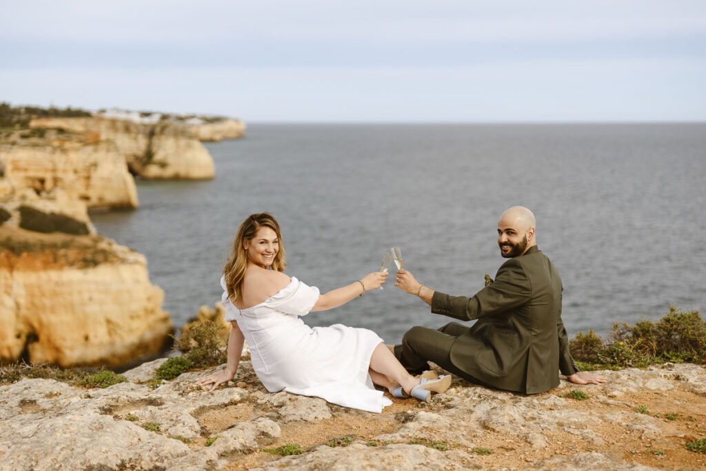 Bride and groom clink champagne glasses on cliffside in Alvor, Portugal