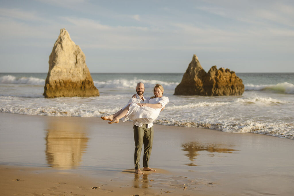 Groom holds bride in arms on beach in Alvor, Portugal