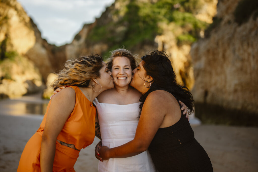 Bridesmaids kiss brides cheek on beach in Alvor, Portugal