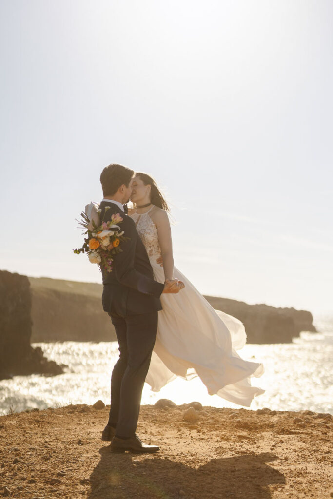 Groom holds bride at cliffside in Bordeira, Portugal
