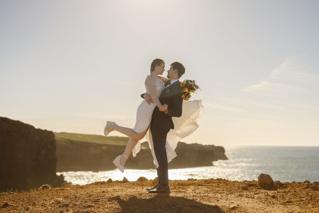 Groom holds bride at cliff in Bordeira, Portugal