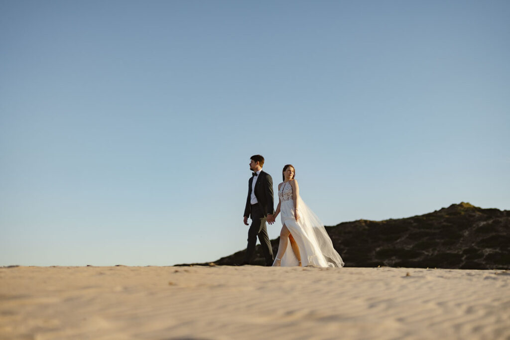 Bride and groom walk down beach in Bordeira, Portugal