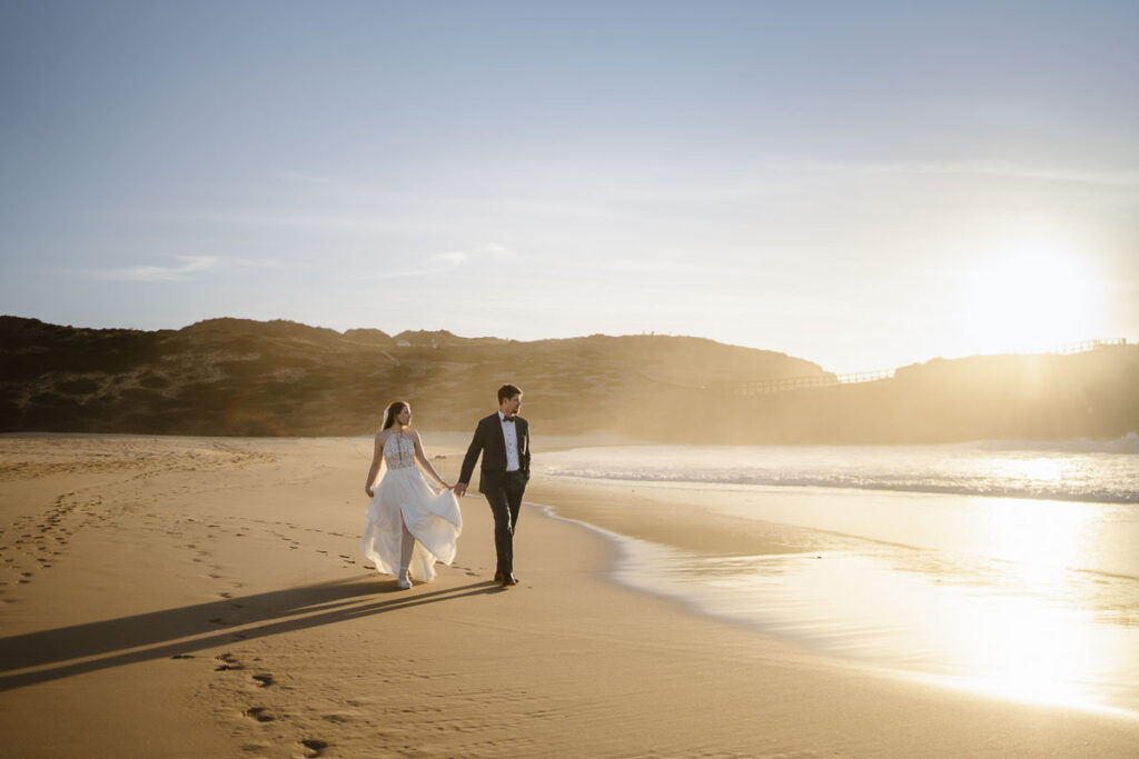 Bride and groom walk down beach in Bordeira, Portugal